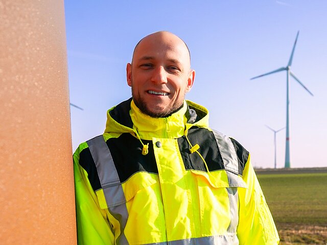 JUWI Mitarbeiter lächelt in die Kamera. Im Hintergrund stehen Windkraftanlagen.