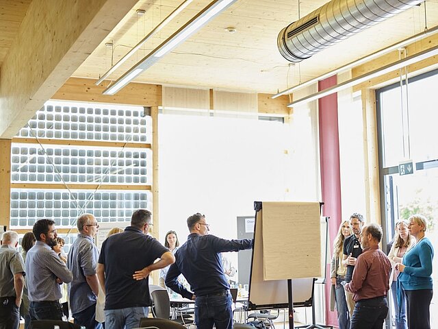 Eine Gruppe von Menschen steht um ein Flipchart und hört den Erklärungen einer weiteren Person zu.